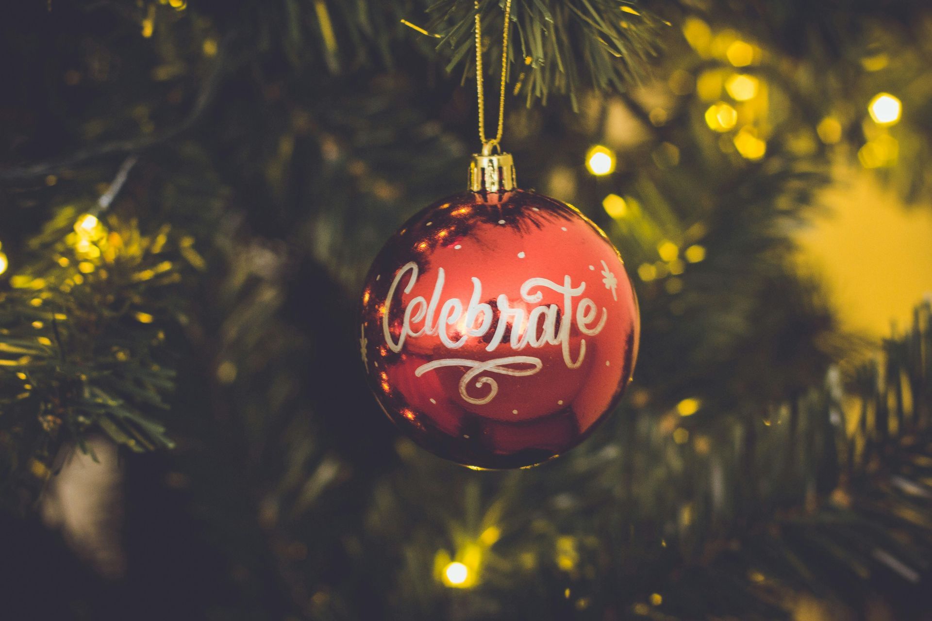 Red Christmas ornament with "Celebrate" in white script, hanging on a tree with warm lights.
