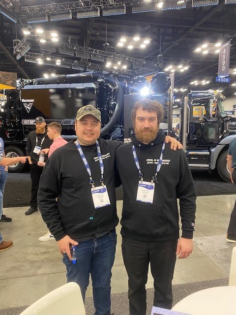 Two men with lanyards pose in front of a large truck indoors.