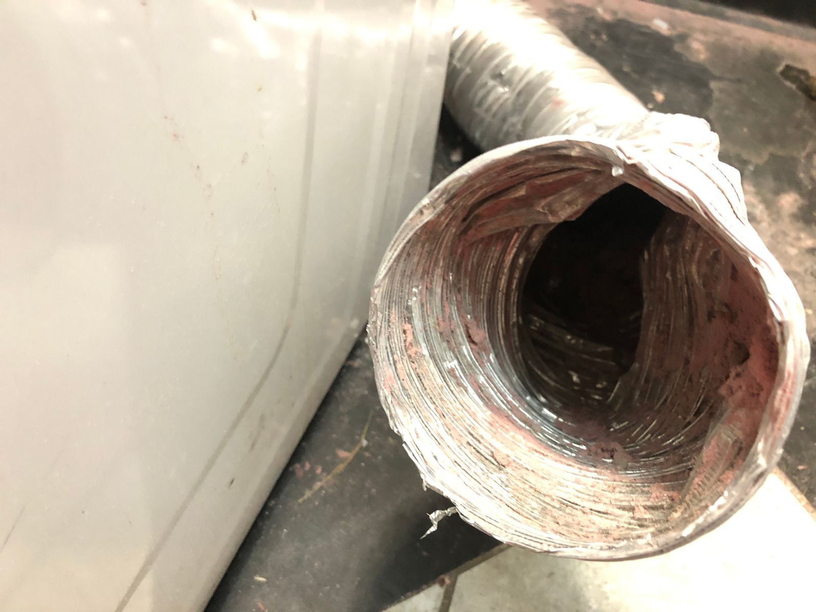 Close-up of a damaged dryer vent tube with a white appliance to the left and a dark floor below.