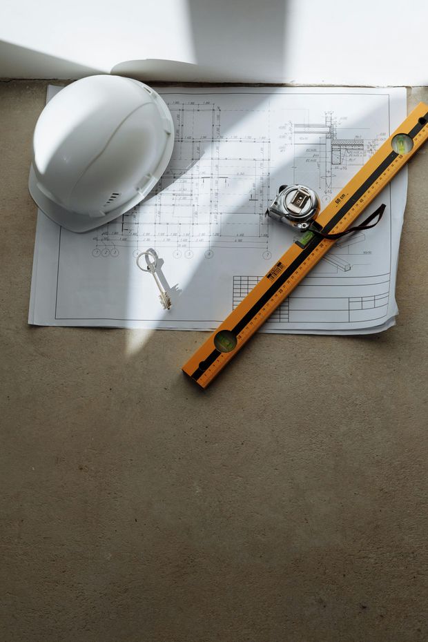 White hard hat, blueprints, key, tape measure, and yellow level on a light brown surface.
