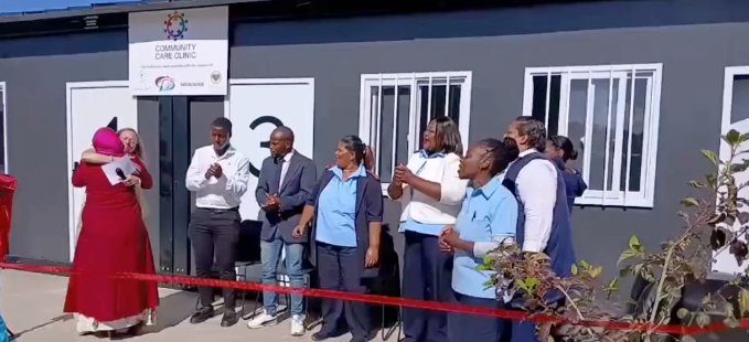 People gathered outside a building, singing near a red ribbon during a ceremony.