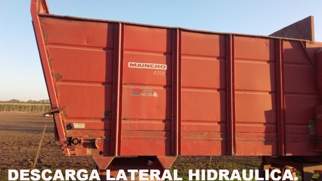 Remolque agrícola rojo con descarga lateral hidráulica en el campo, con las palabras “DESCARGA LATERAL HIDRAULICA”.