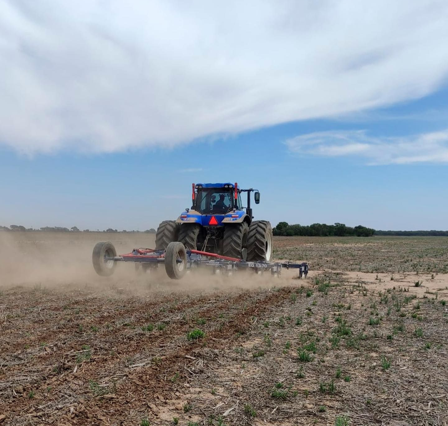 Tractor azul arando un campo, levantando polvo bajo un cielo parcialmente nublado.