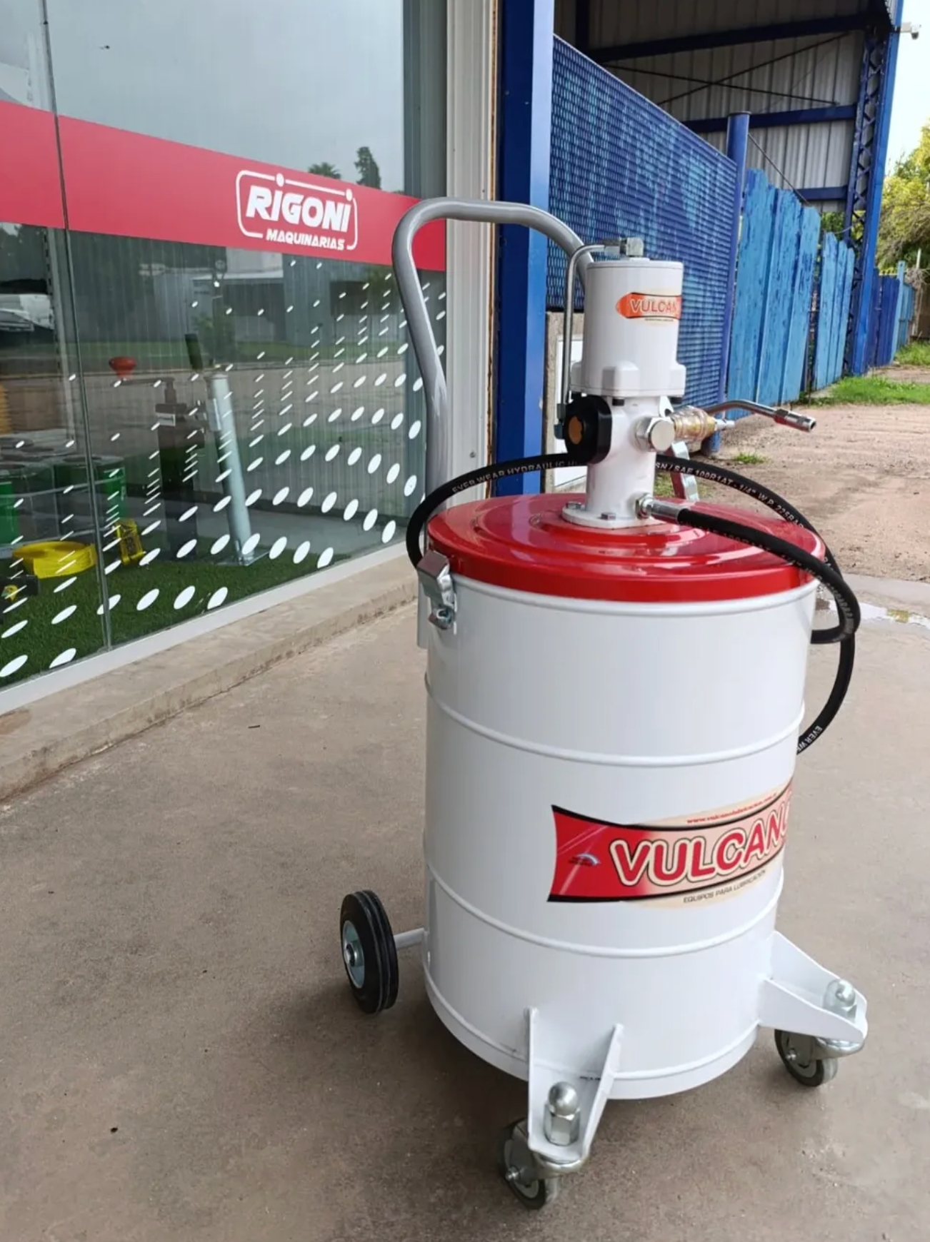 Bomba de grasa VULCAN blanca y roja sobre ruedas con asa, junto a una vitrina de cristal.