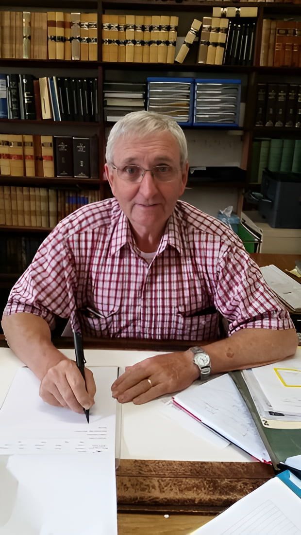A Man is Sitting at a Desk in Front of a Bookshelf Writing on a Piece of Paper — Hynes & McCormack In Murwillumbah, NSW
