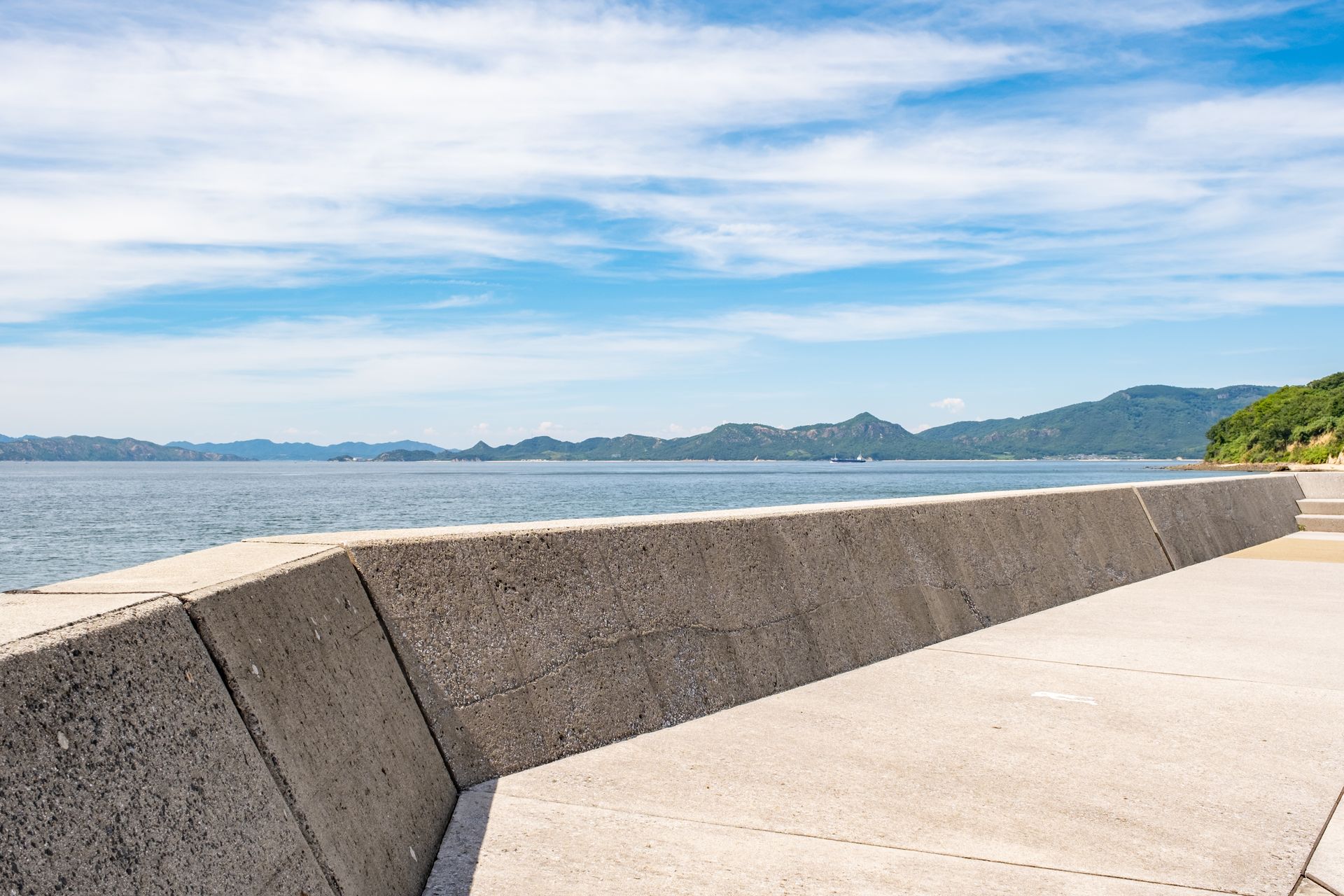 Concrete seawall along a calm sea under a blue sky with some clouds, mountains in the distance.