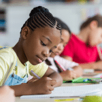 A group of children are sitting at desks in a classroom writing in notebooks.