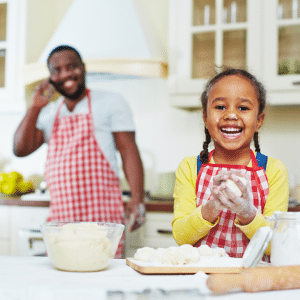 A little girl is smiling in a kitchen while a man talks on a cell phone.