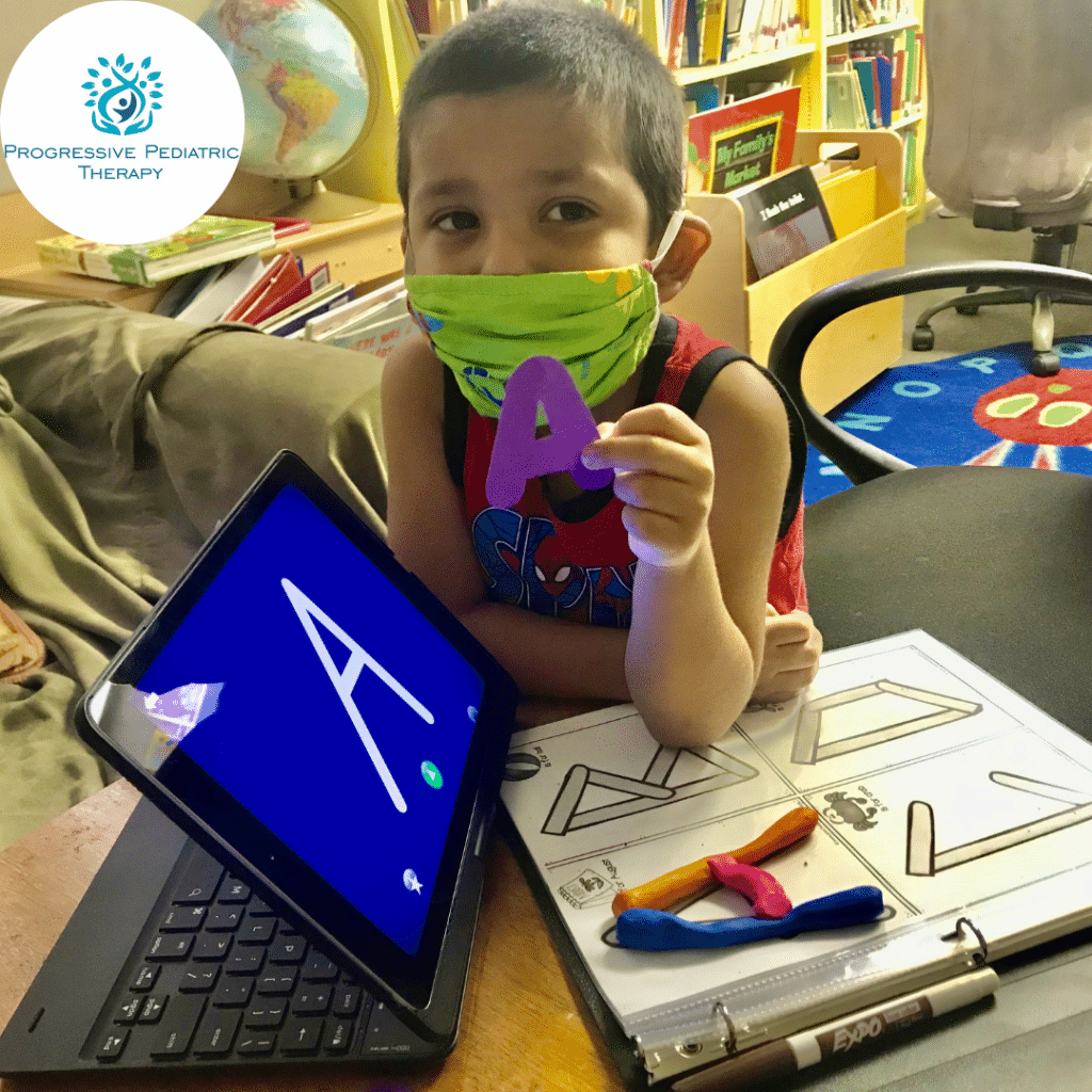A young boy wearing a mask is holding a letter a in front of a laptop.