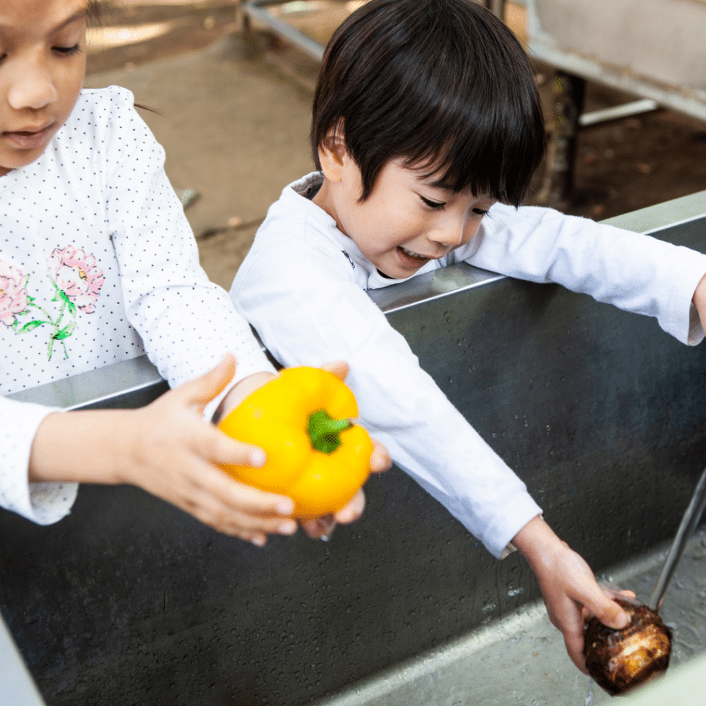 Two children are washing vegetables in a sink and one is holding a yellow pepper