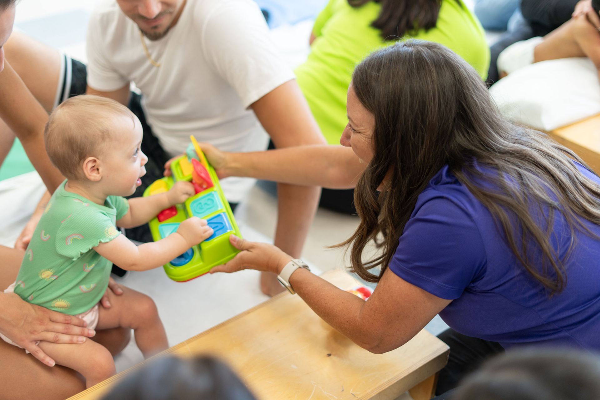 Baby reaching for a toy held by a woman, surrounded by others, playing indoors.