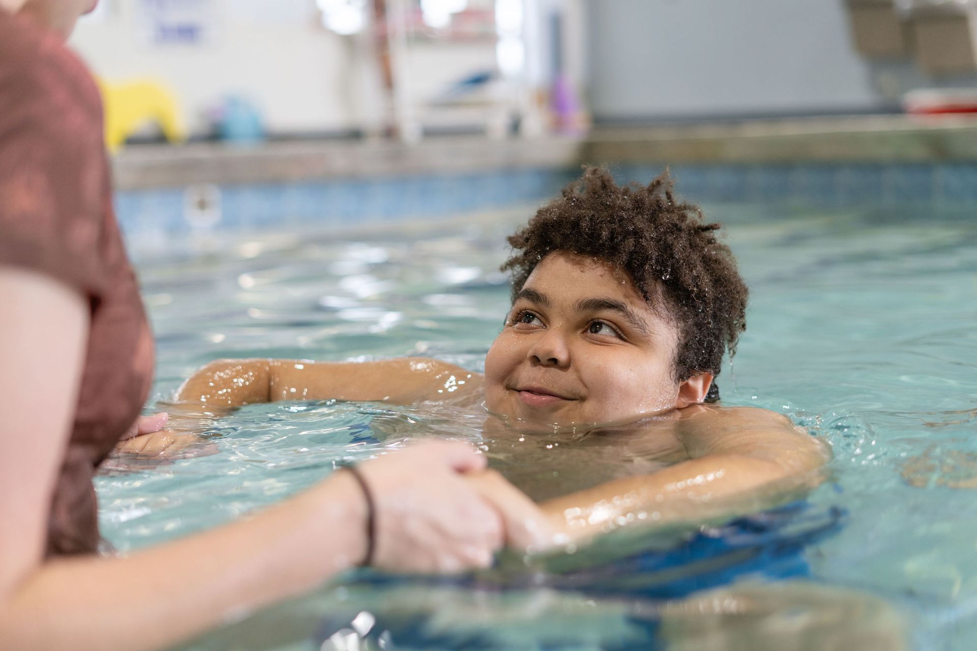 A young man is swimming in a swimming pool while a woman holds his hand.