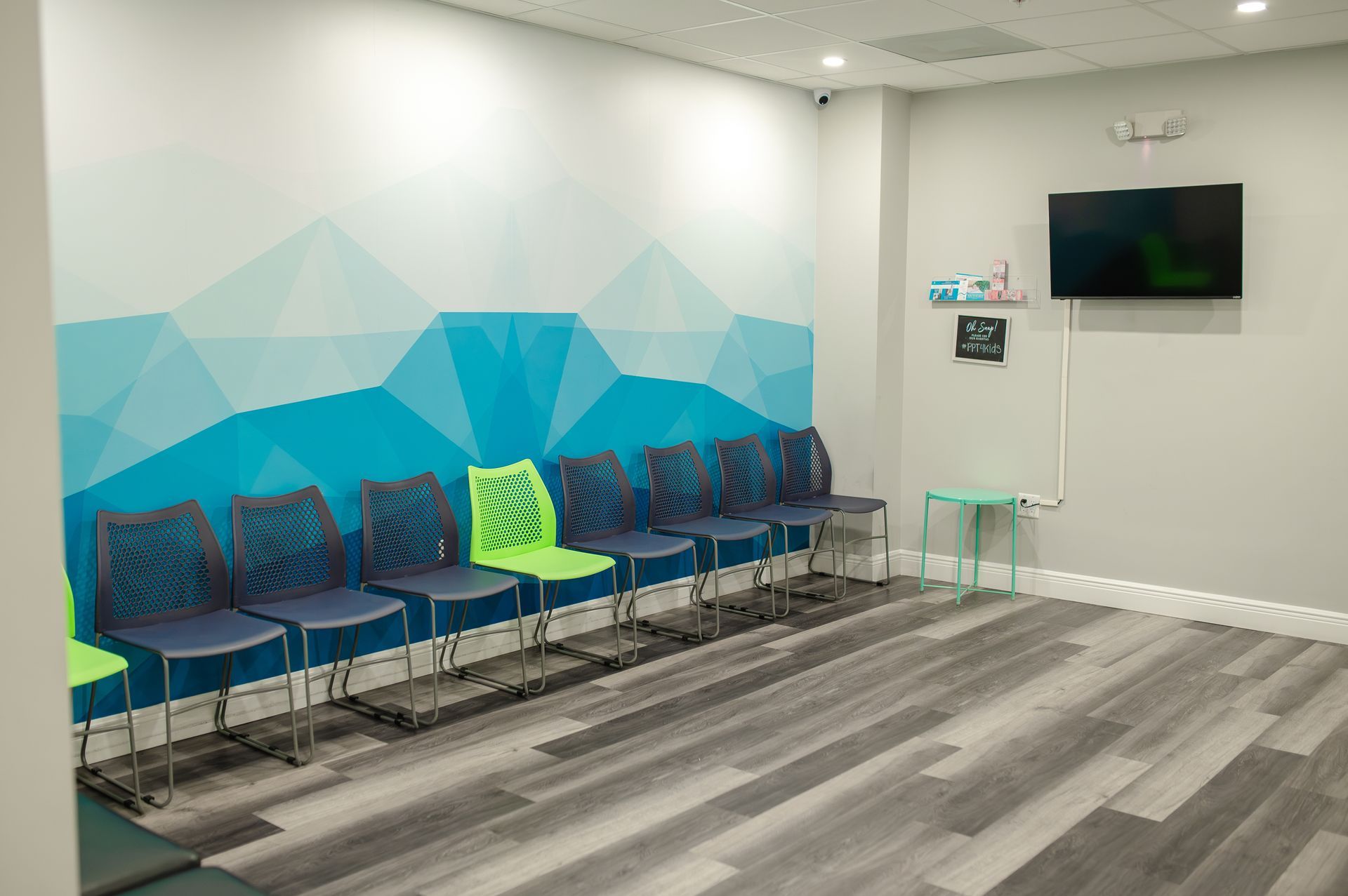 Waiting room with blue and green chairs, geometric wall art, television, and light gray flooring.
