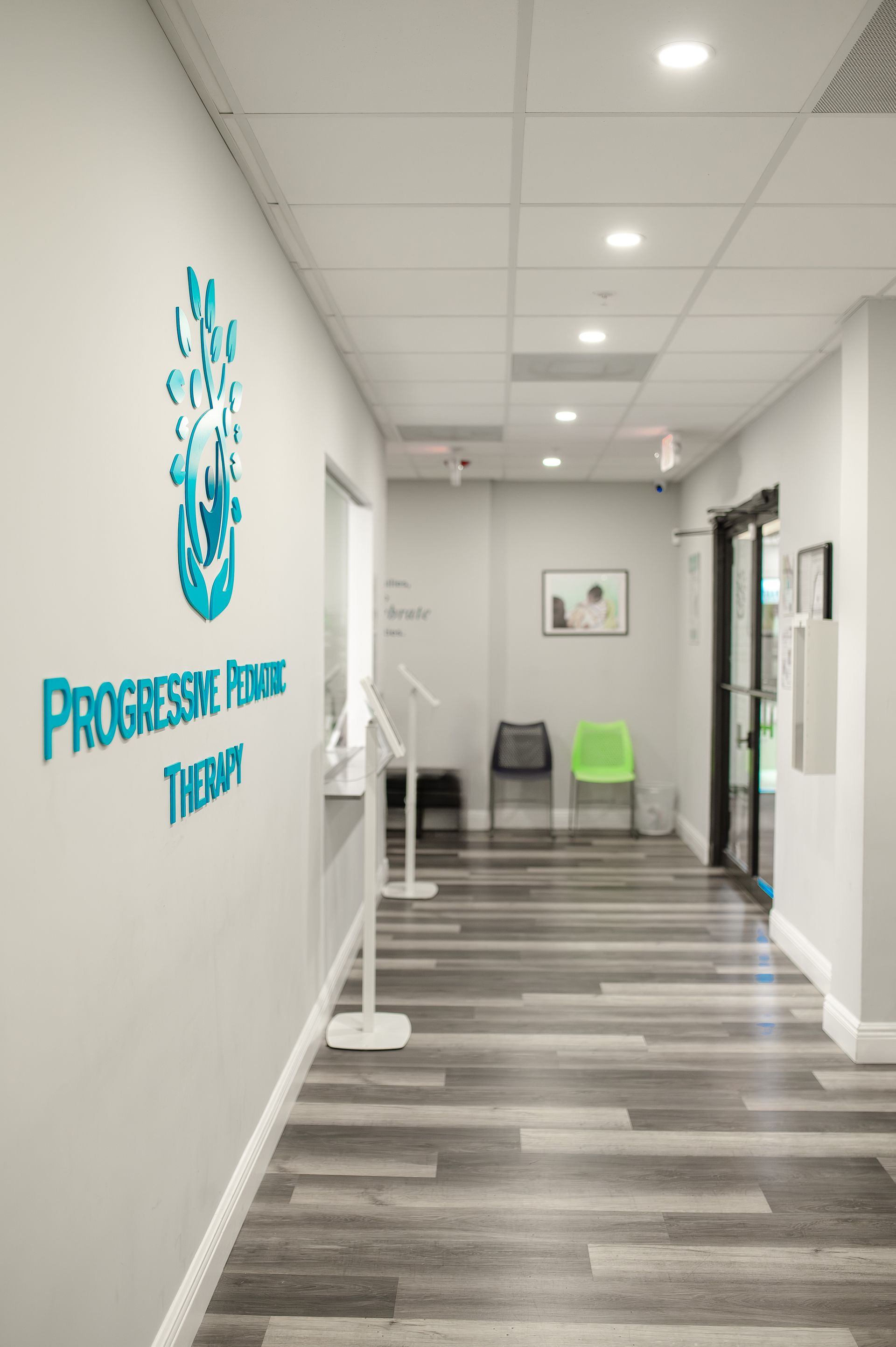 Hallway of a physical therapy clinic with logo on wall, waiting area with chairs, and a door at the end.