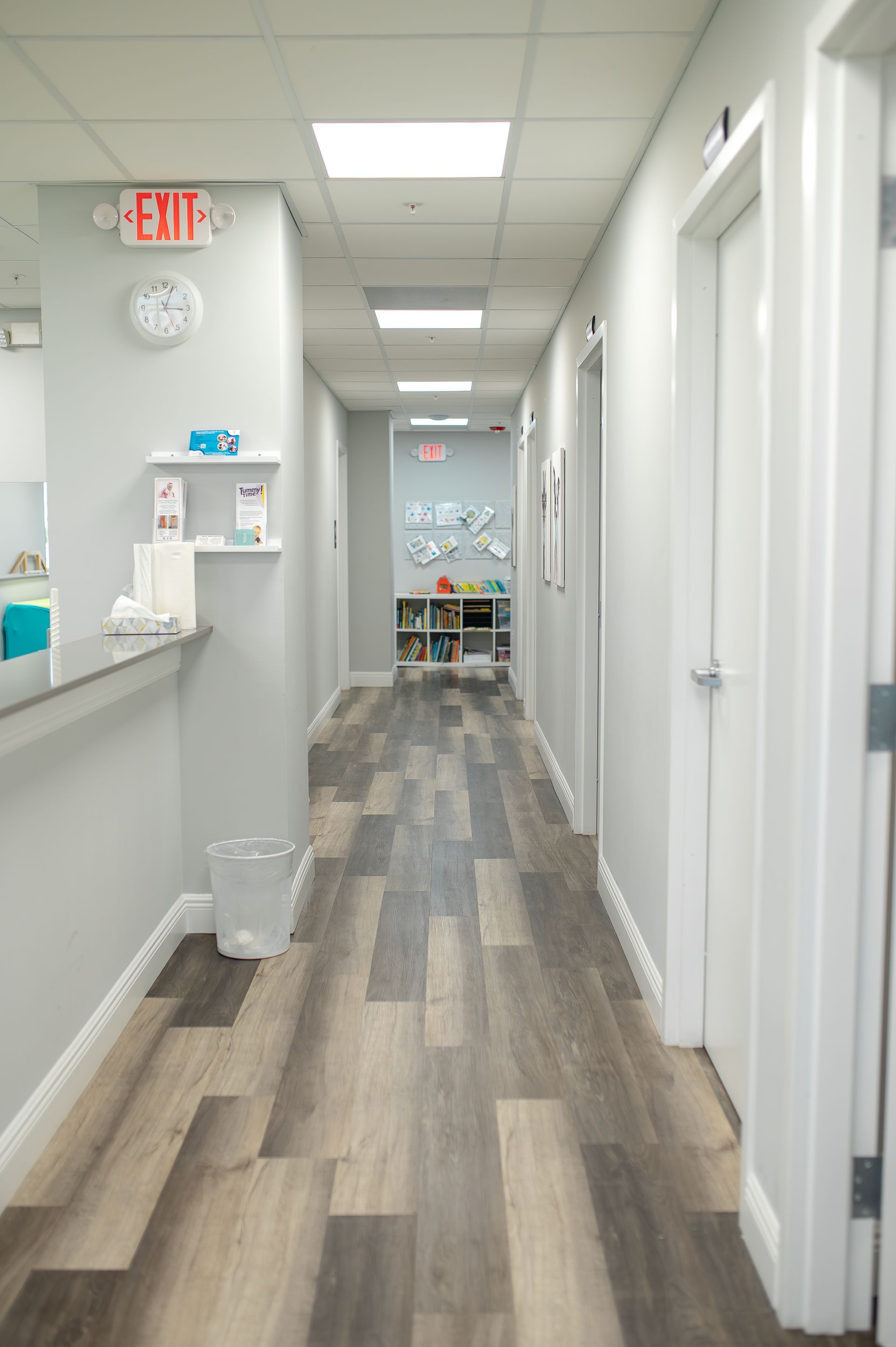 Hallway with gray walls, wood-look flooring, and white doors. An exit sign and clock are visible.