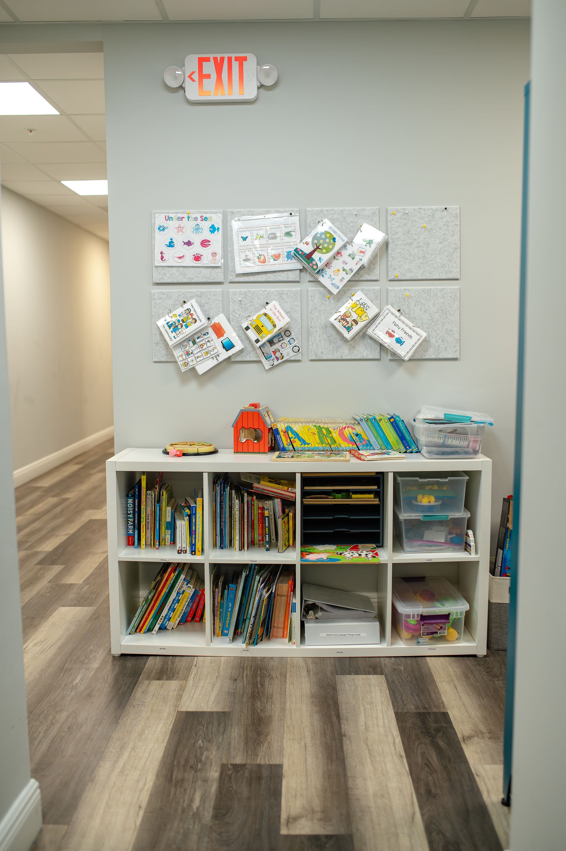 Hallway with a white shelf containing books and art supplies; artwork on a corkboard. 