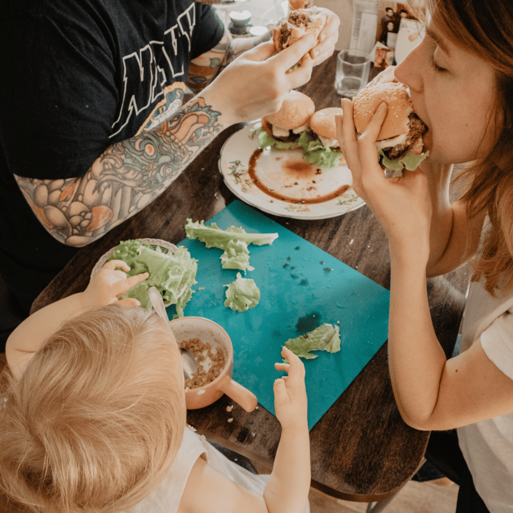 A man in a navy shirt is eating a hamburger with a woman and a baby.