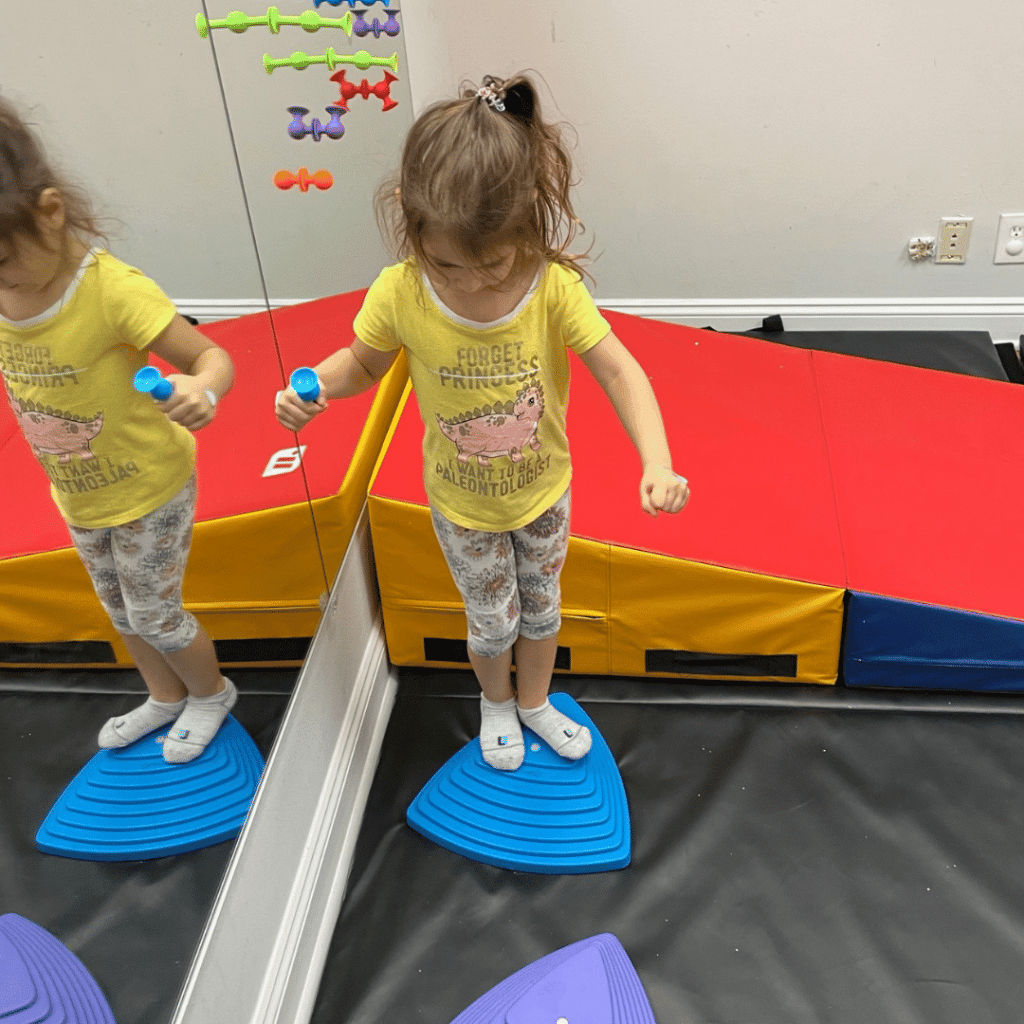 A little girl in a yellow shirt is standing on a blue platform in front of a mirror.