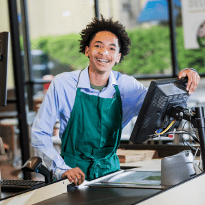 A man in a green apron is standing in front of a computer.