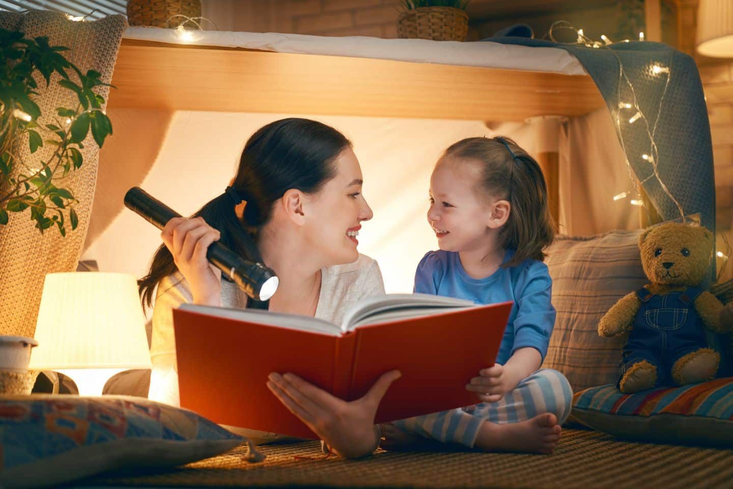 A woman is reading a book to a little girl while holding a flashlight.