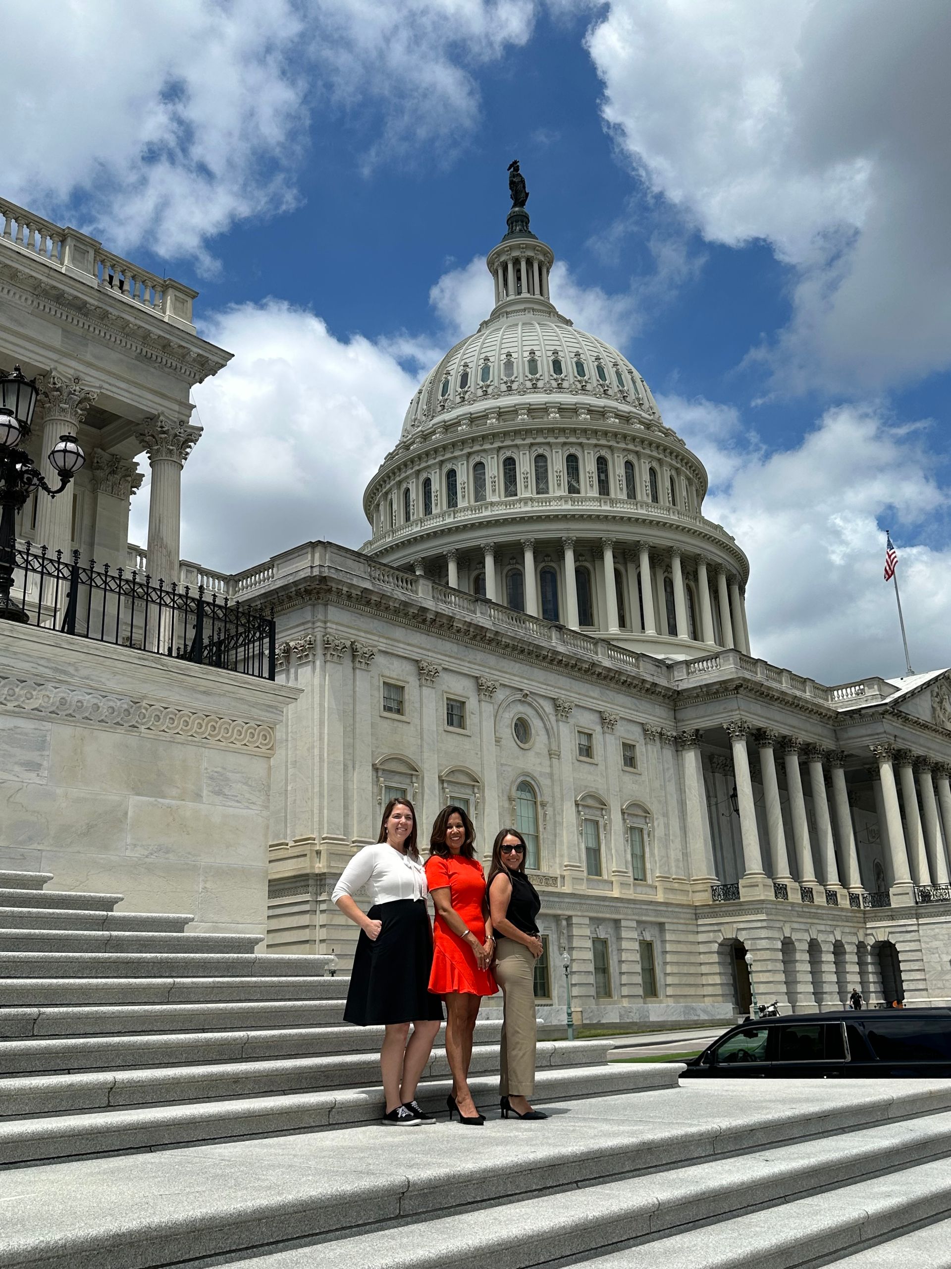 Three women pose on steps in front of the U.S. Capitol Building on a sunny day.