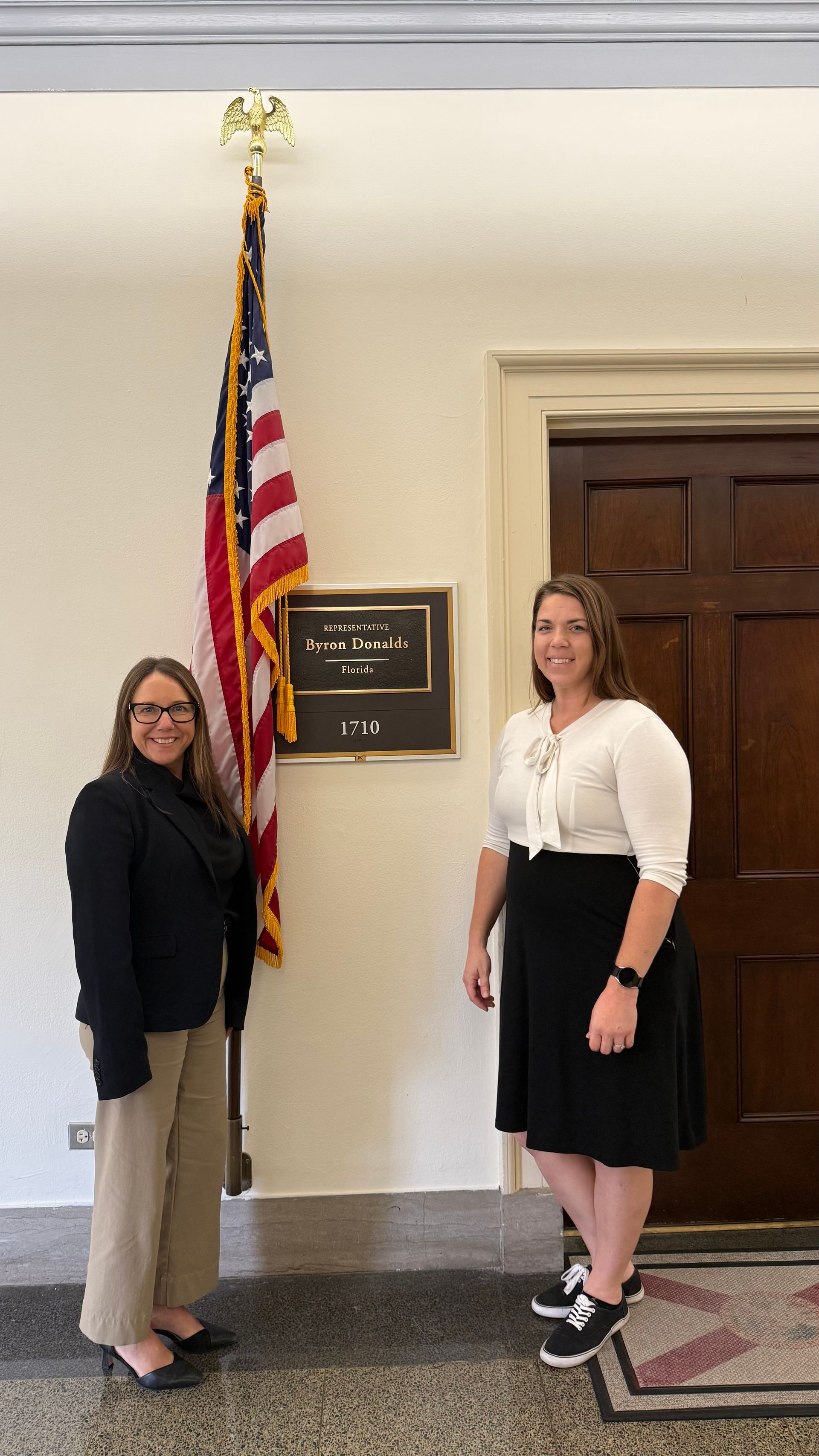 Two women standing near an American flag and plaque in a hallway. One wears a blazer, the other a white top and black skirt.