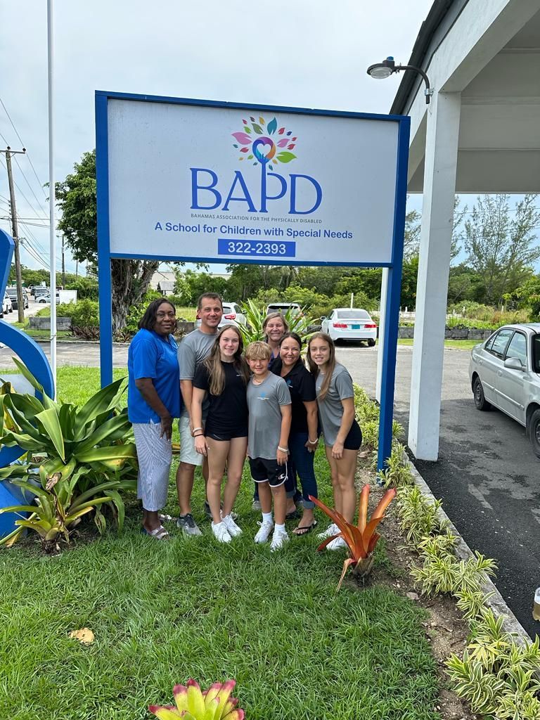 Group of people in front of a blue sign for BAPD. Lush greenery and cars in the background.