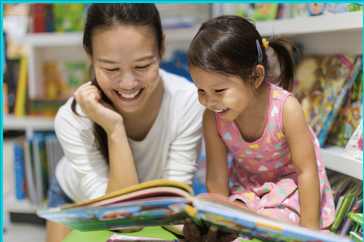 A woman and a little girl are reading a book together in a library.