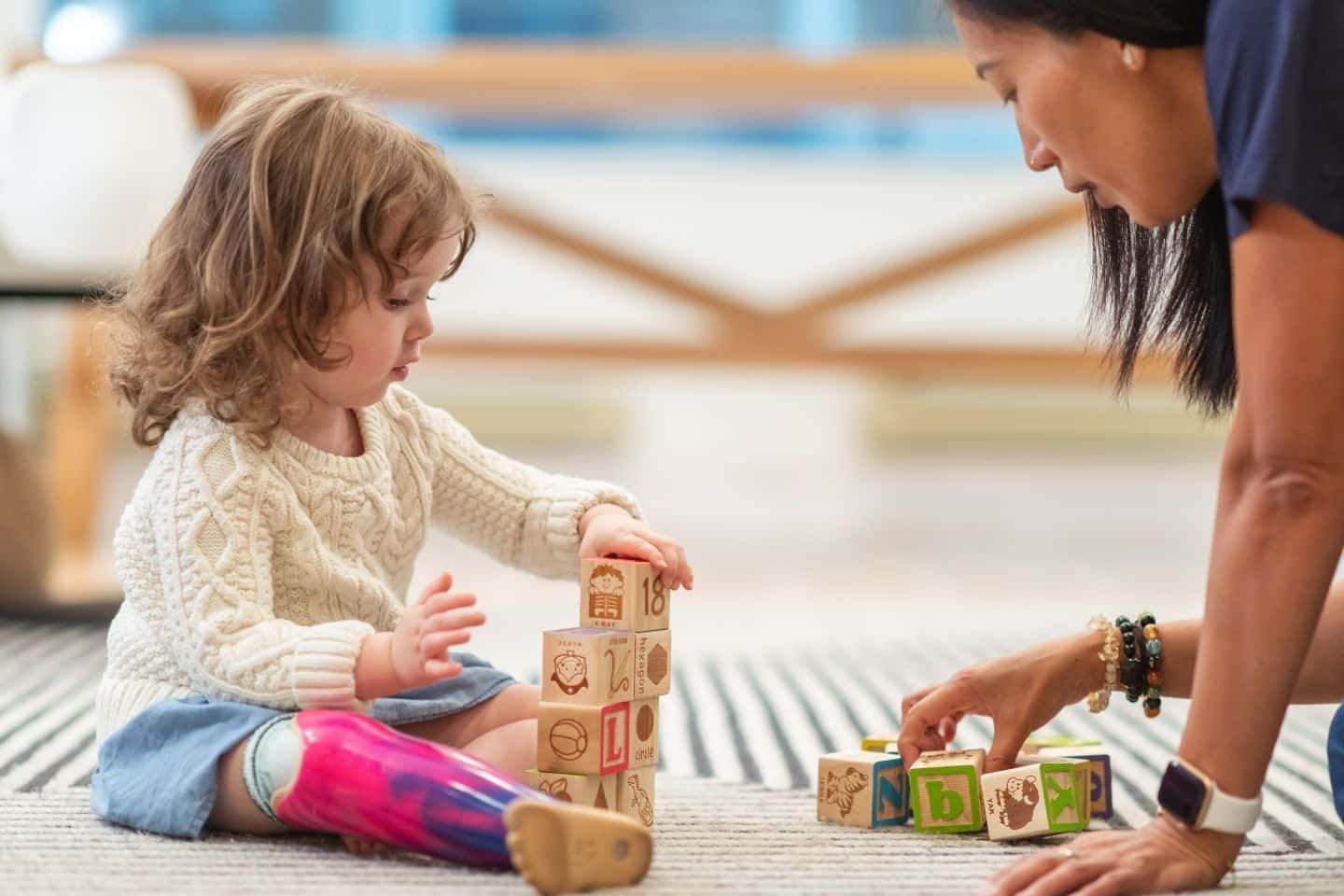 A woman and a little girl are playing with wooden blocks on the floor.