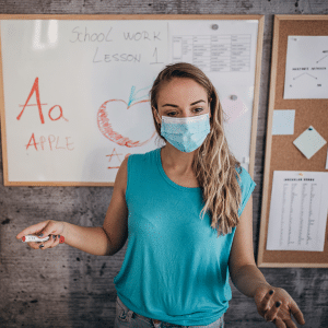 A woman wearing a mask is standing in front of a whiteboard that says school work lesson 1