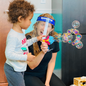 A woman wearing a face shield is playing with a child who is blowing soap bubbles