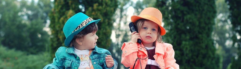 Two little girls are sitting next to each other in a park talking on a cell phone.