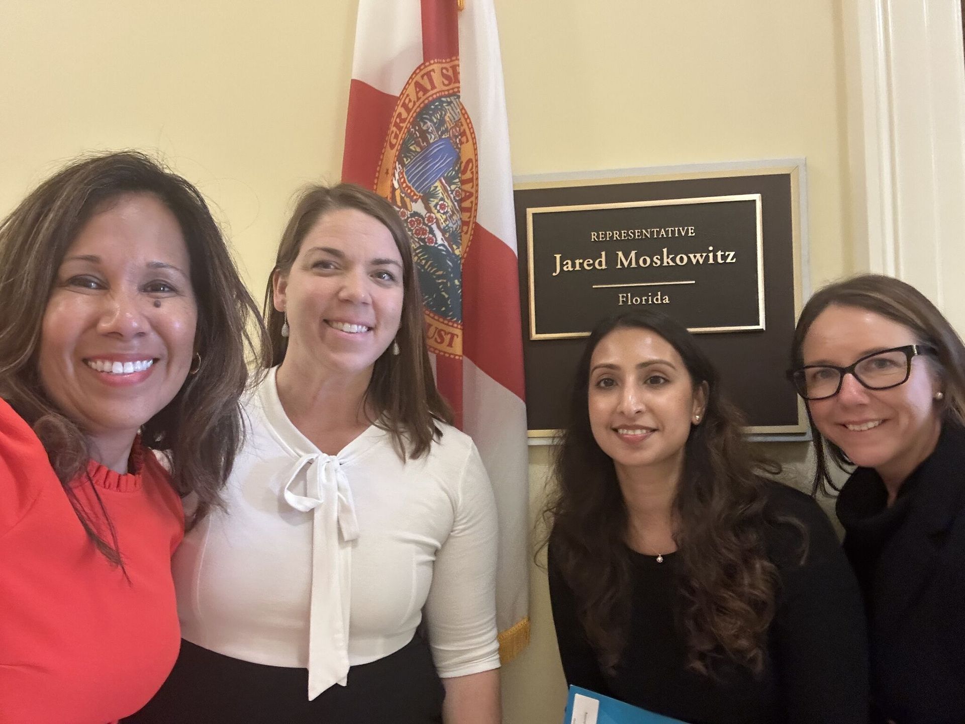 Four women smiling for a photo in front of a Florida flag and sign for Jared Moskowitz's office.
