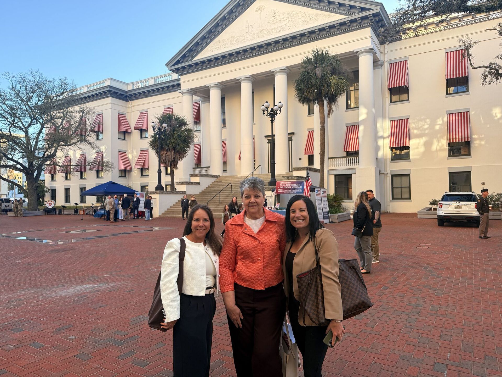 Three women pose in front of a large building with columns. Red and white banners hang from the windows.