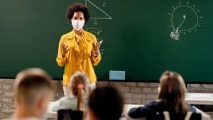 A teacher wearing a mask is teaching a classroom of children.