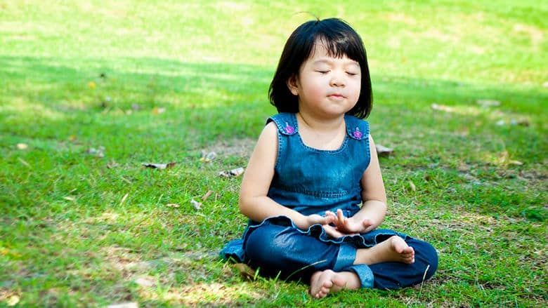 A little girl is sitting in a lotus position on the grass with her eyes closed.