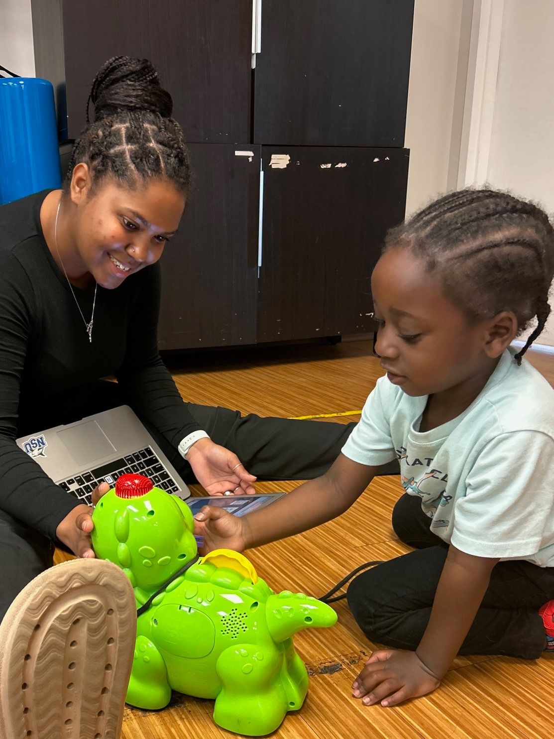 A woman and a child are playing with a toy dinosaur on the floor.
