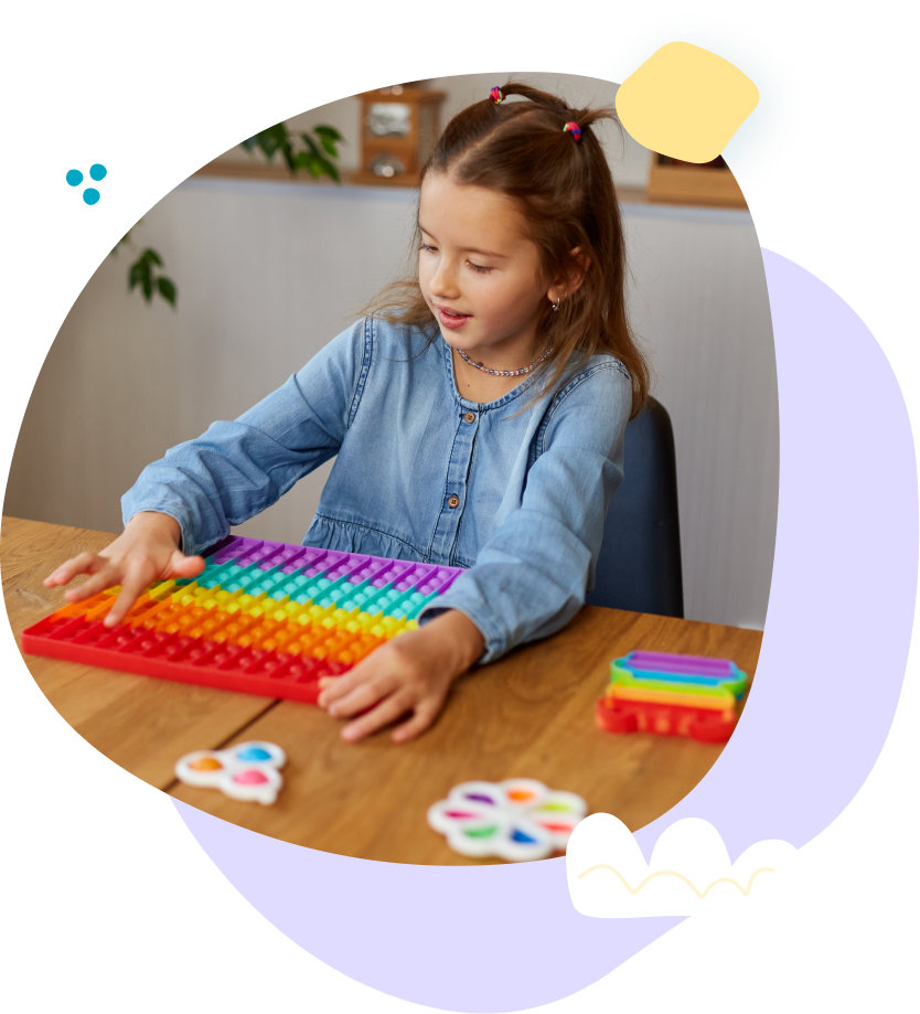 A little girl is sitting at a table playing with a rainbow pop it toy.