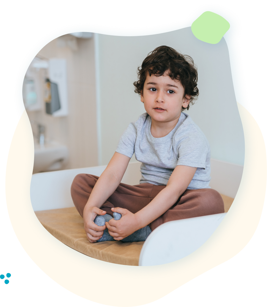 Boy sitting cross-legged, looking forward. Wearing gray shirt and brown pants. Indoors, natural light.