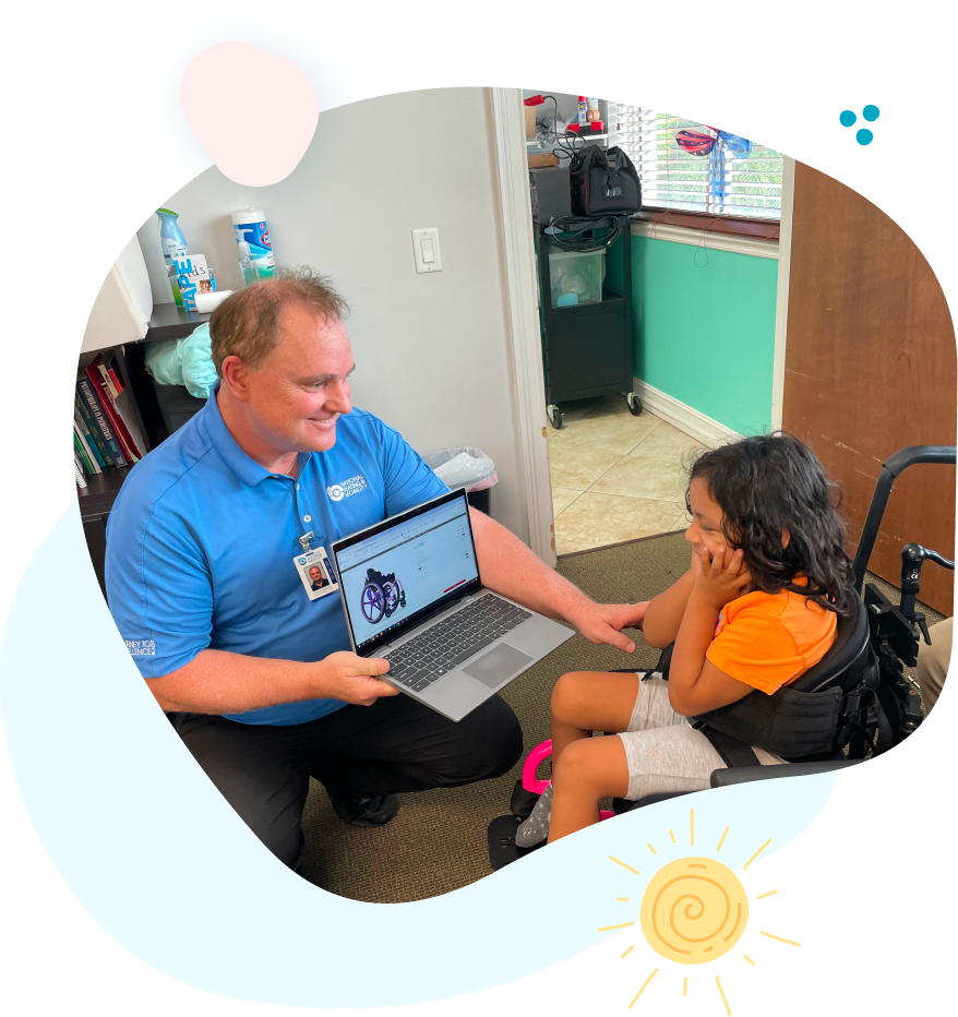 A man is kneeling down next to a girl in a wheelchair while holding a laptop.