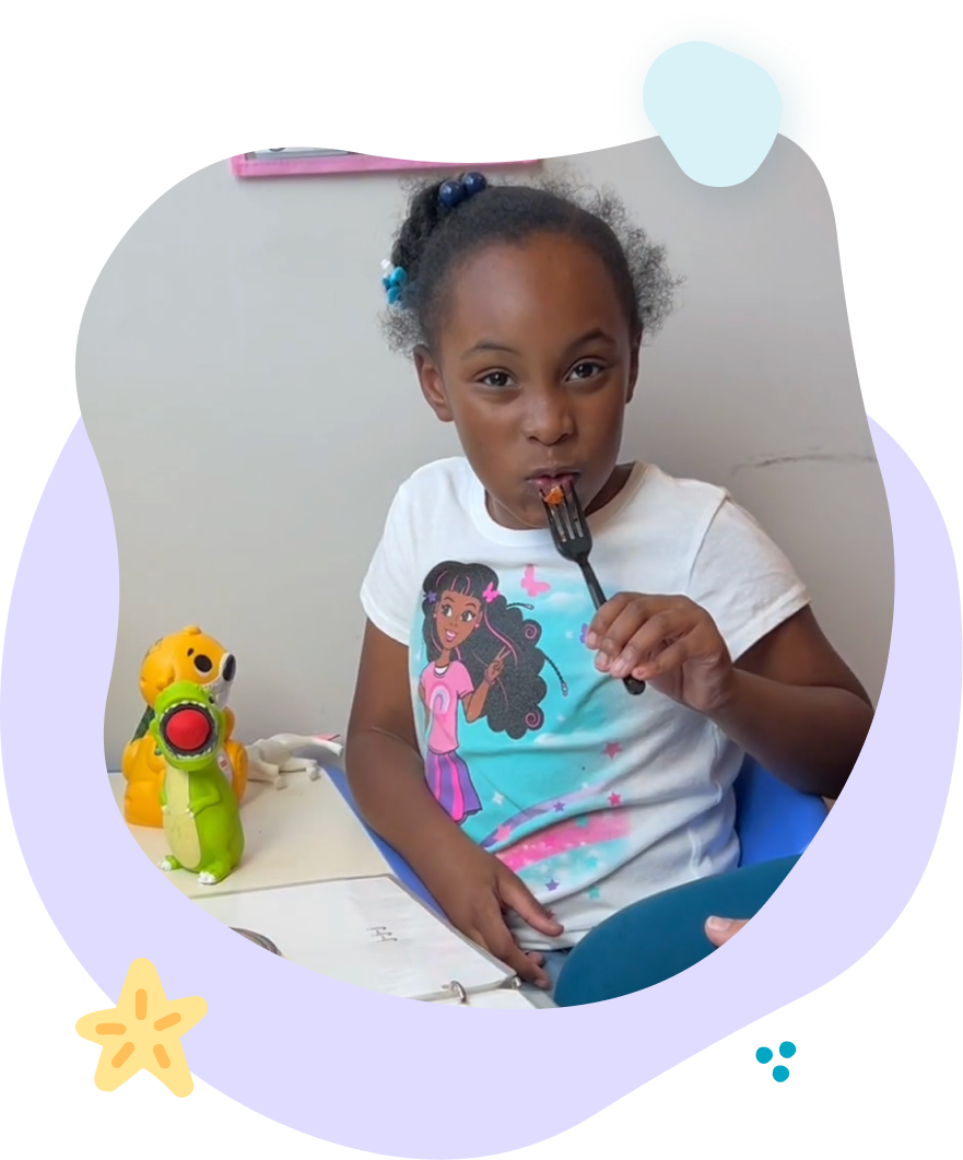 Girl eating from a fork, seated at a table. Wearing a white t-shirt and blue chair. Toys on the table.