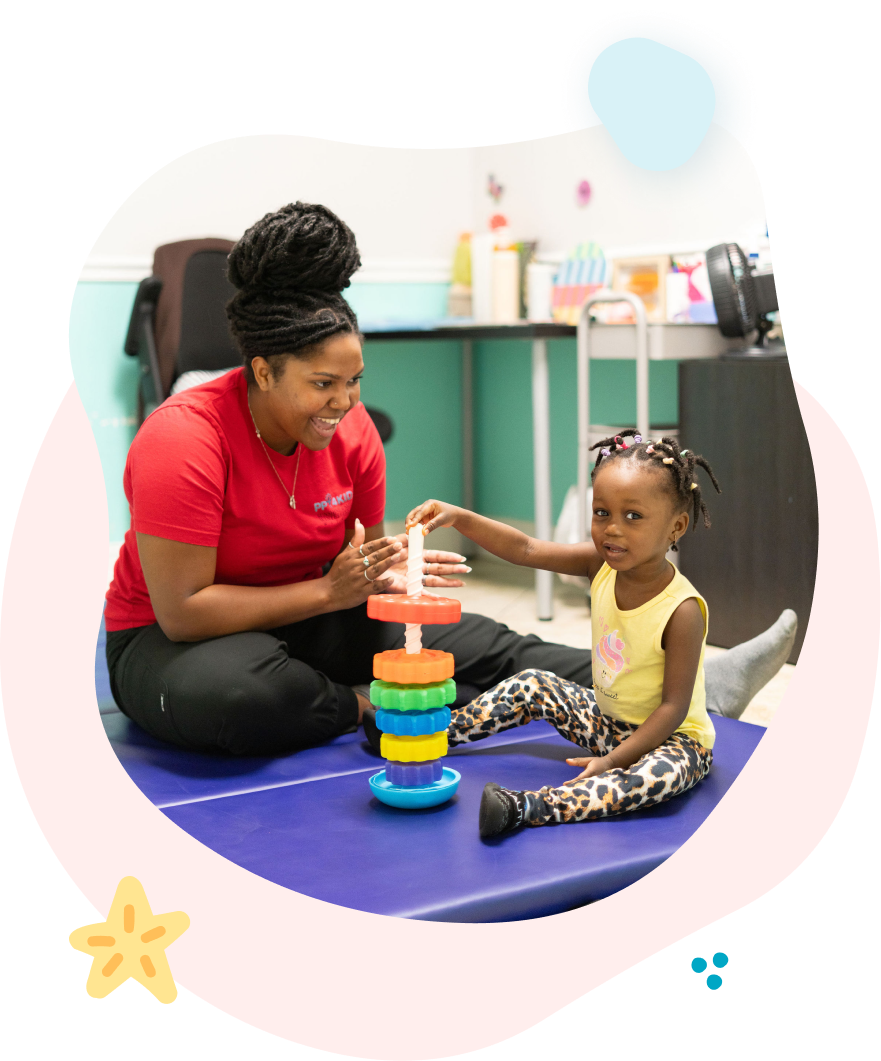 A person in a red shirt helps a child stack rings on a toy tower, both smiling on a floor mat.