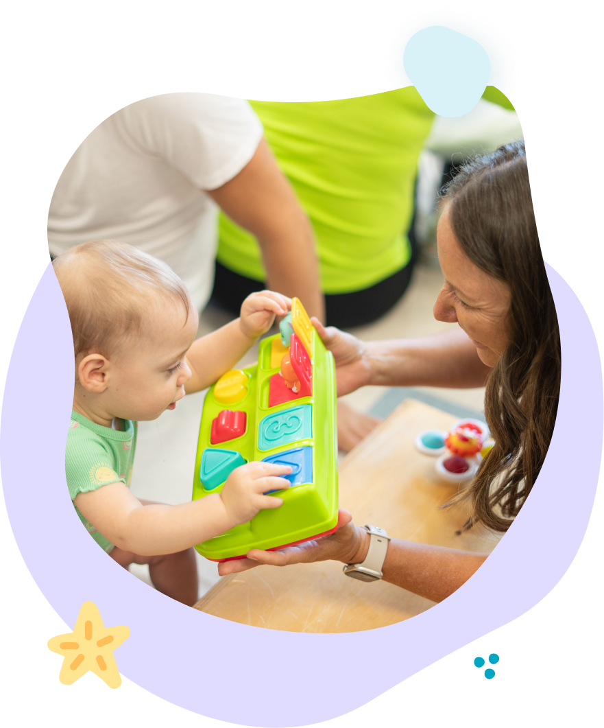 Baby reaching for a colorful toy held by an adult; another person is in the background.