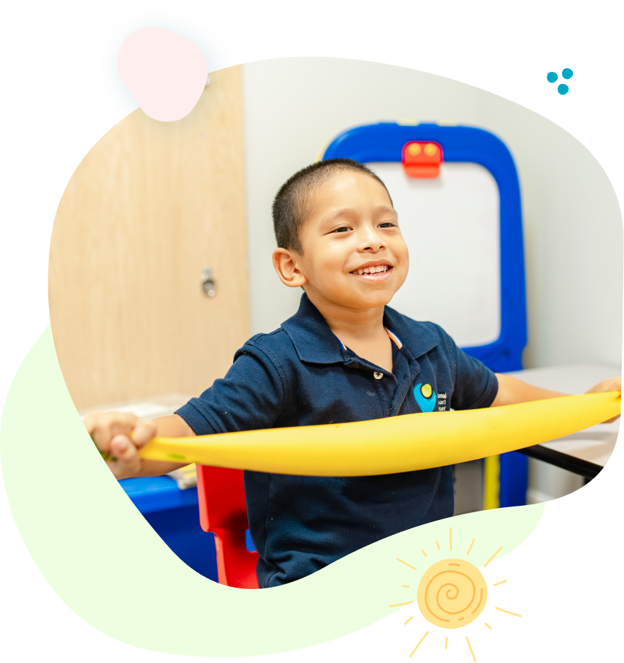 Boy smiles, holding yellow exercise bar in a classroom setting with whiteboard in the background.