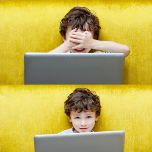 A young boy is covering his eyes while using a laptop computer.