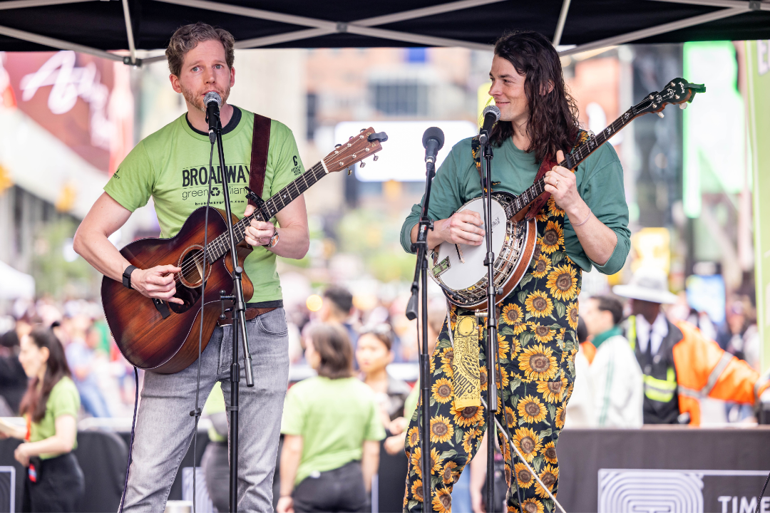 Two musicians performing with a guitar and banjo on the Broadway Celebrates Earth Day stage, wearing Broadway Green Alliance apparel amidst the towering architecture of Midtown Manhattan.