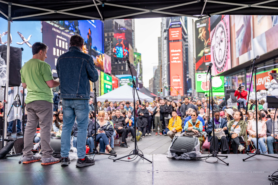 A wide-angle view from behind the performers on stage looking out at a massive crowd gathered in Duffy Square, surrounded by the iconic skyscrapers and digital billboards of the world’s most famous business and entertainment hub.