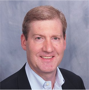 A smiling man with light brown hair wearing a dark blazer over a light-colored button-down shirt against a grey backdrop.