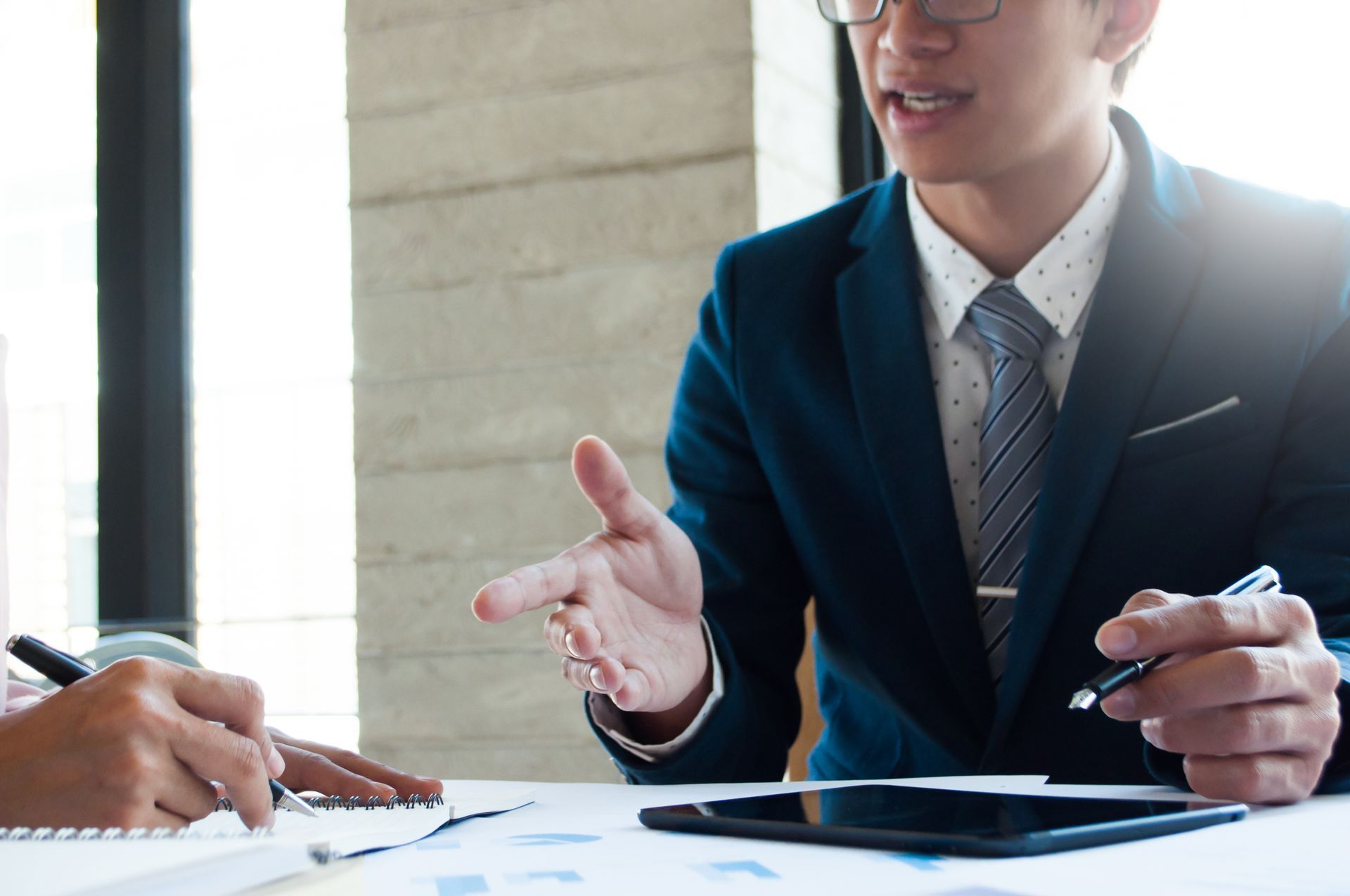 Two people in professional attire discuss work over a tablet and documents at a desk.
