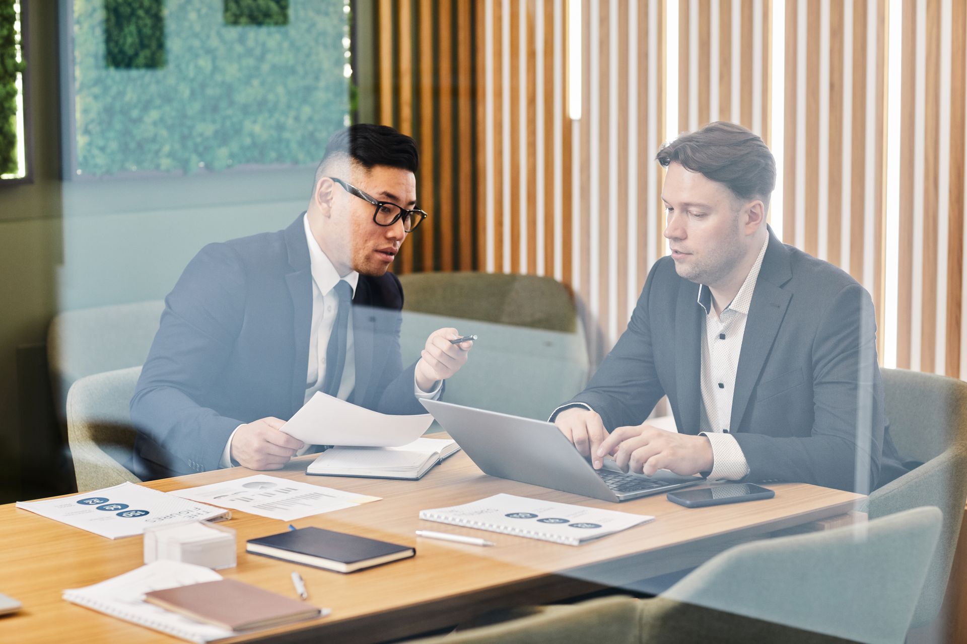 Two colleagues in suits collaborate in a modern office, one typing on a laptop and the other reviewing a document.